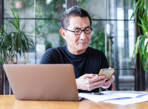 man with glasses sitting at a desk looking at his mobile phone with a laptop off to the side